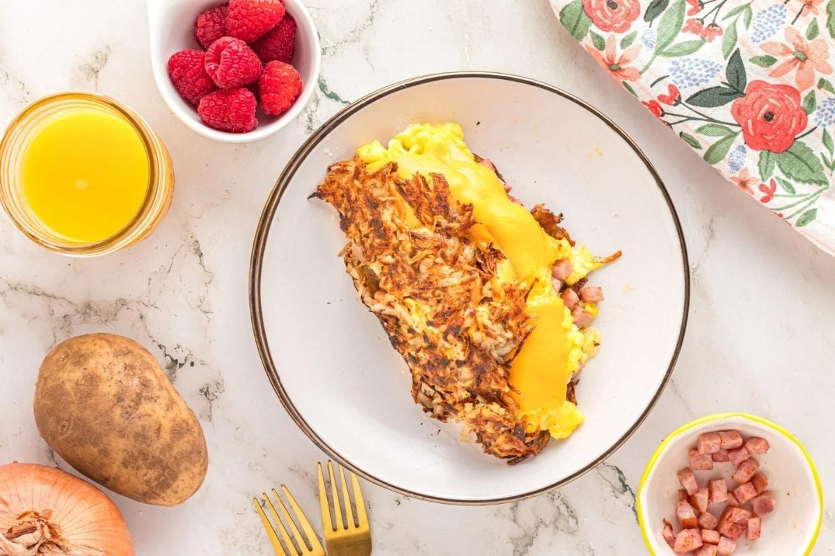 A plate of omelette with melted cheese and hash browns served with a side of raspberries and a glass of orange juice, perfect for Father's Day breakfast. Ingredients like onion, potato,