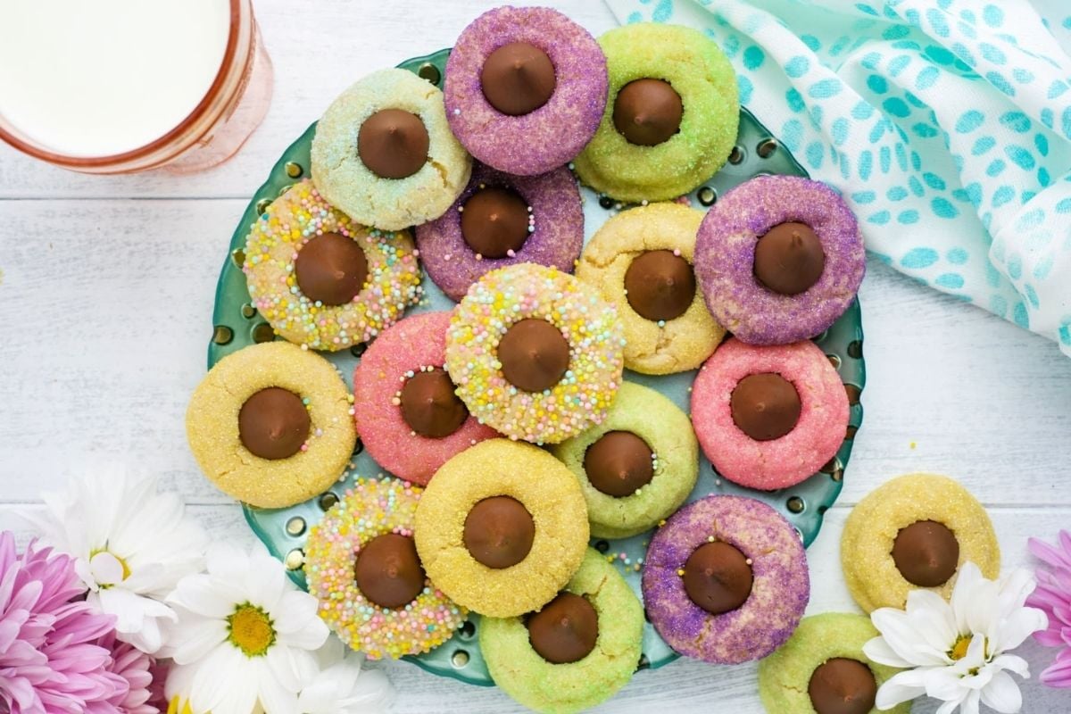 A plate of colorful ring-shaped cookies with chocolate centers, accompanied by a glass of milk and surrounded by flowers, is featured in our cookie recipes collection.