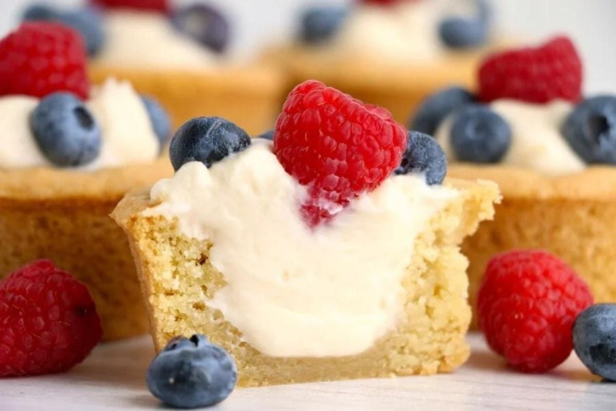A mini cheesecake topped with a dollop of cream and fresh berries, featuring a cookie crust, with a bite taken out of it to reveal the creamy filling.