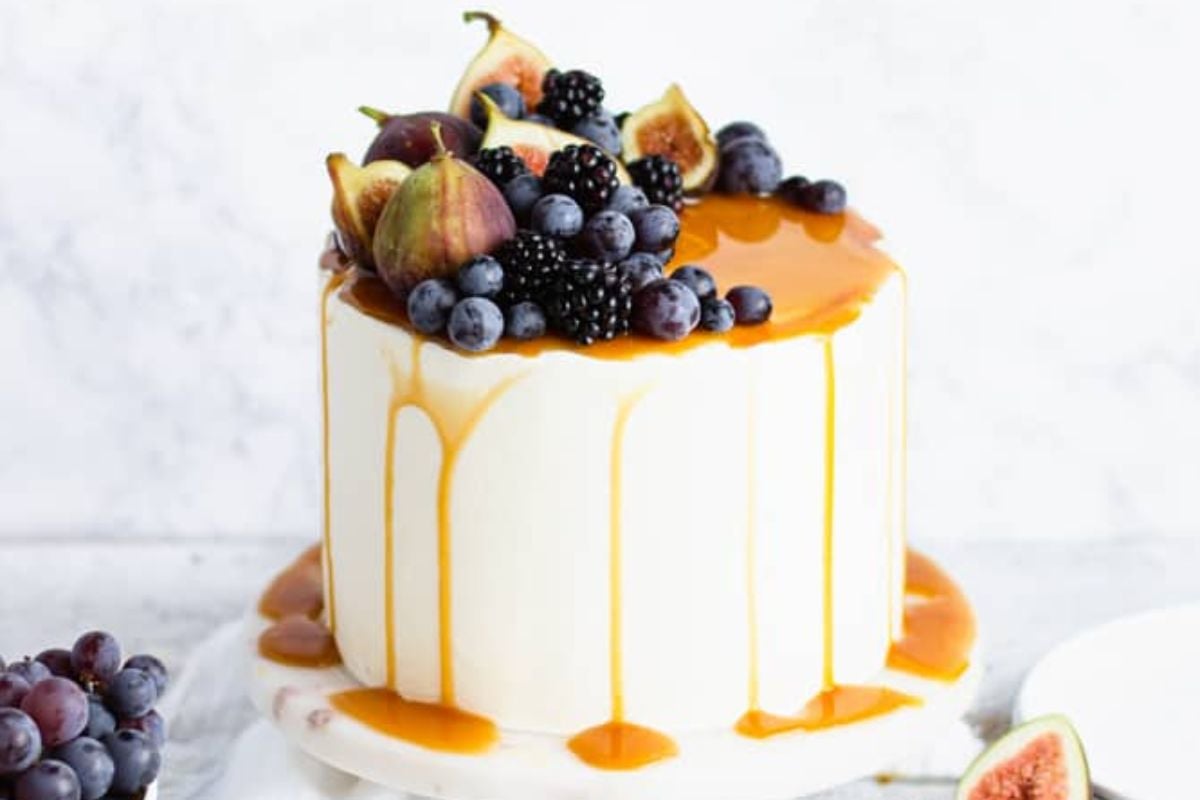 A layer cake with white frosting, caramel dripping down the sides, and topped with assorted fresh fruit including figs and berries.