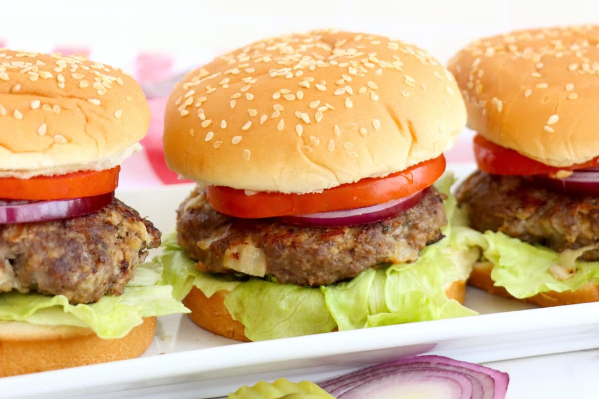 Three hamburgers with lettuce, tomatoes, and onions on sesame seed buns, inspired by old-fashioned recipes.