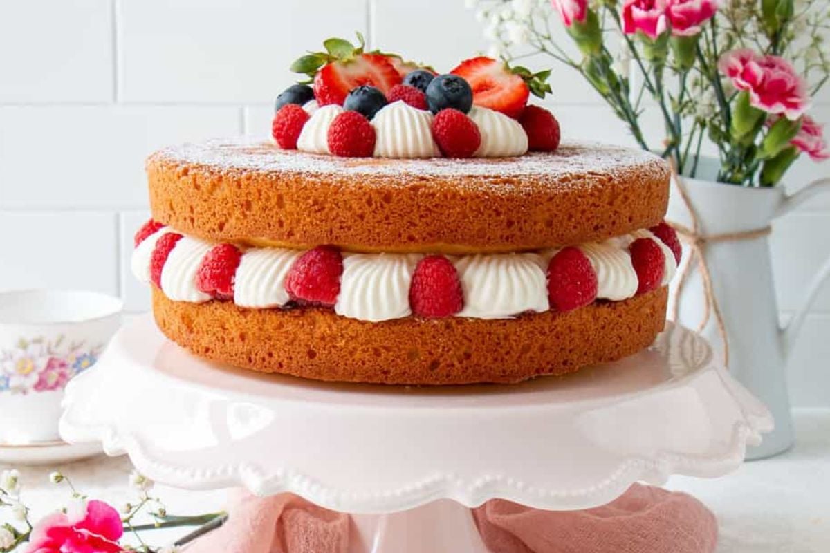 A two-layer sponge cake with cream and fresh berries on a pink cake stand, with layer cakes and flowers in the background.