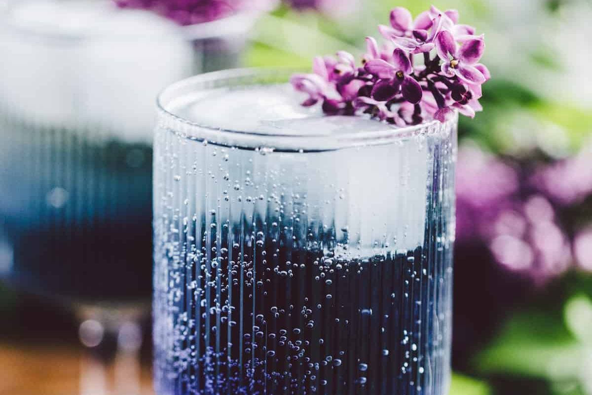 A close-up of a glass with a blue Easter cocktail garnished with purple flowers, with more glasses in the background.