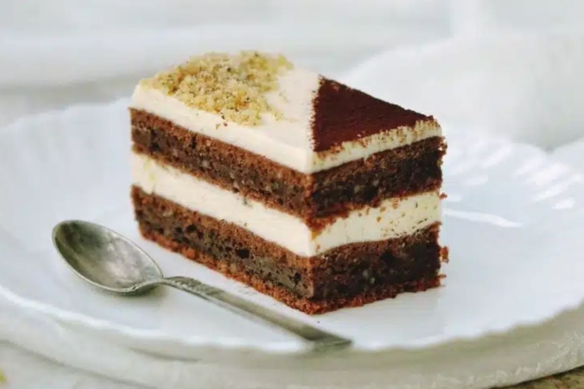 A slice of layered chocolate and layer cakes with cream on a white plate with a spoon to the side.