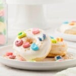 Delightful donuts with white icing and colorful marshmallows sit on plates beside a jar brimming with the vibrant charms of Lucky Charms, all arranged elegantly on a marble surface.