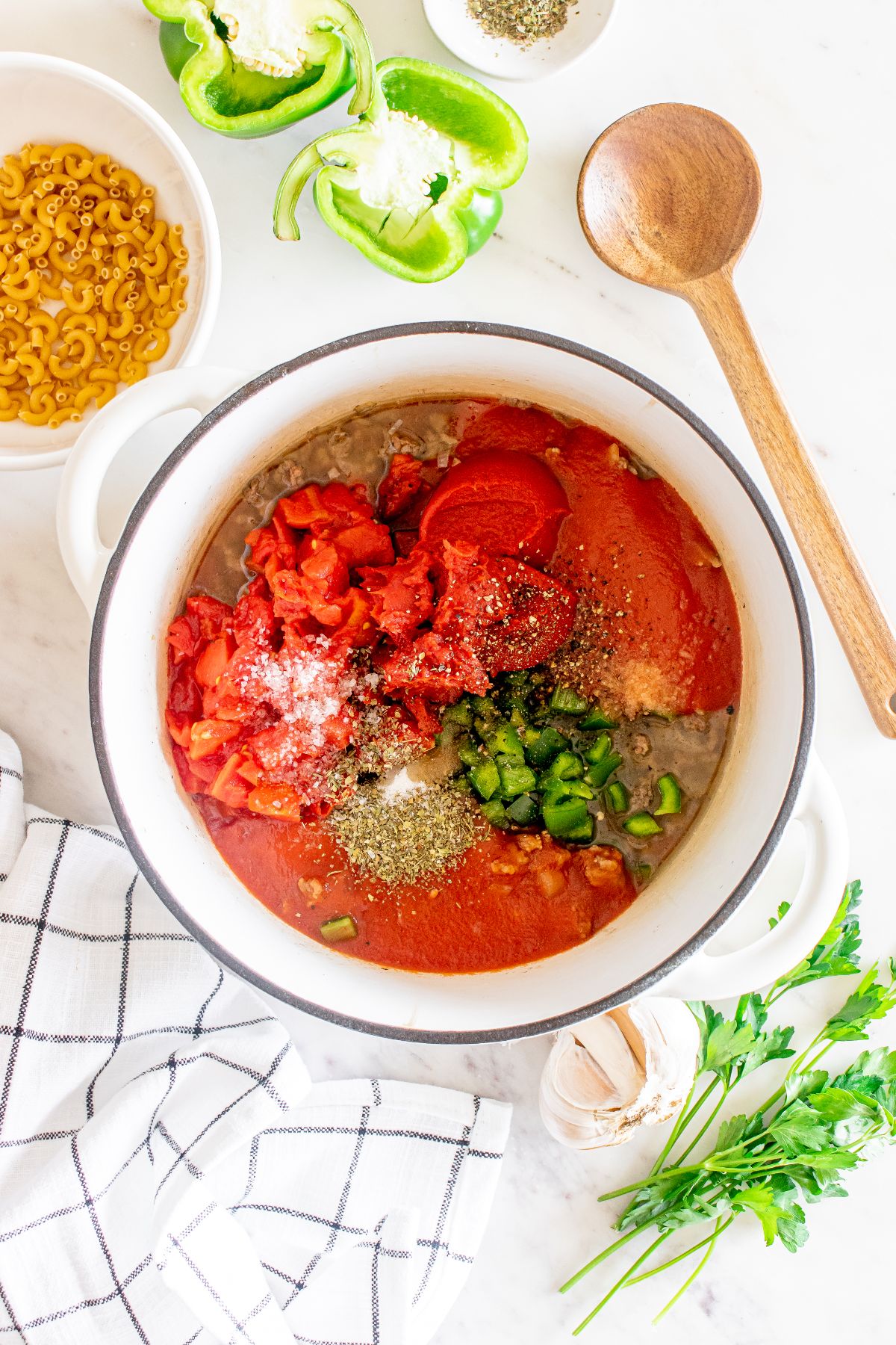 Overhead view of a pot with tomatoes, peppers, and seasonings—classic American chop suey ingredients—surrounded by pasta, veggies, and a wooden spoon.