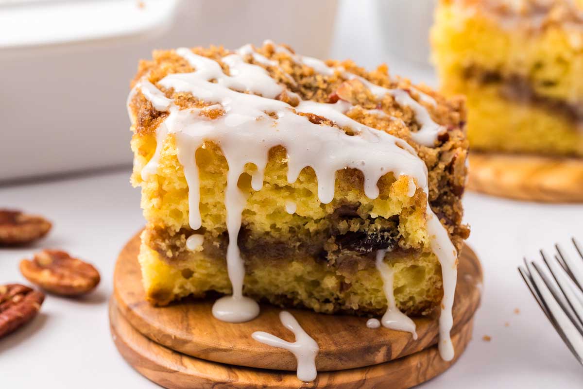 A close-up of a slice of cinnamon coffee cake with layers of brown sugar and pecan filling, topped with white icing. The cake sits on a wooden coaster, and a fork is placed nearby. Pecans are scattered in the background.