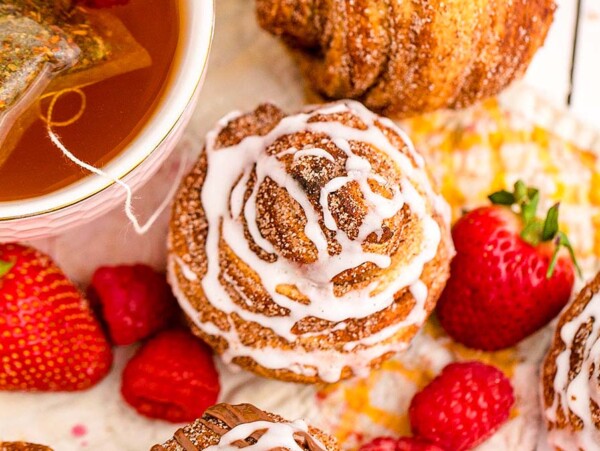 A cup of tea with a tea bag, cinnamon pastries with icing, strawberries, and raspberries on a white surface—perfect inspiration for your next brunch recipes.