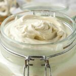 A glass jar filled with creamy white substance, possibly homemade whipped shea butter, with herbs in the background.