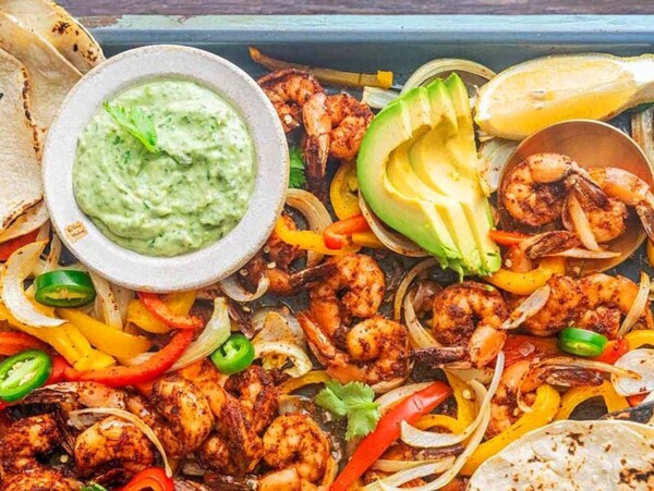 Mexican inspired shrimp fajitas with grilled peppers, onions, avocado, lime, tortillas, and green sauce on a tray.