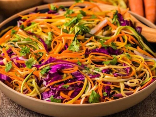 A bowl of fresh vegetable salad with spiralized carrots, cucumber, purple cabbage, and cilantro, featuring delicious additions from our favorite zucchini recipes, with a wooden serving spoon beside it.