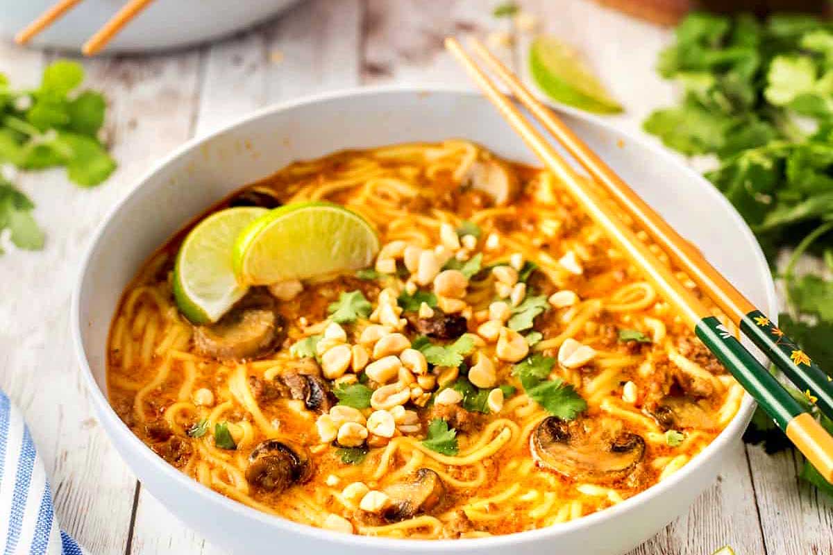 A bowl of spicy noodle soup garnished with lime, peanuts, cilantro, and mushrooms, ready in 30 minutes or less, served with chopsticks on the side.