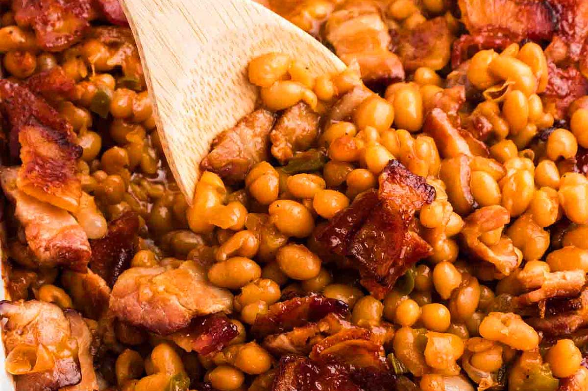 A close-up image of a dish with cooked lentils, chunks of bacon, ketchup, and green peas mixed together, with a wooden spoon.
