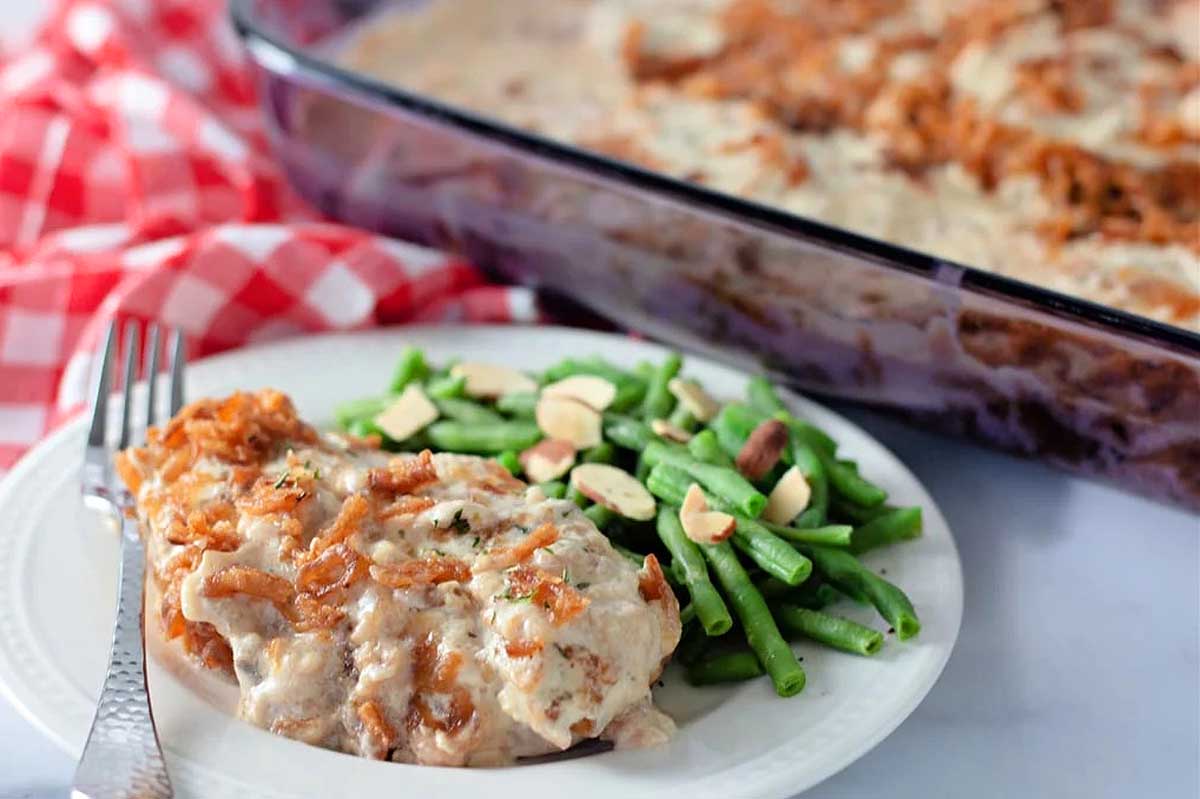 A serving of lasagna on a white plate with green beans and sliced almonds, beside a large glass dish of pork chop recipes on a red checkered tablecloth.