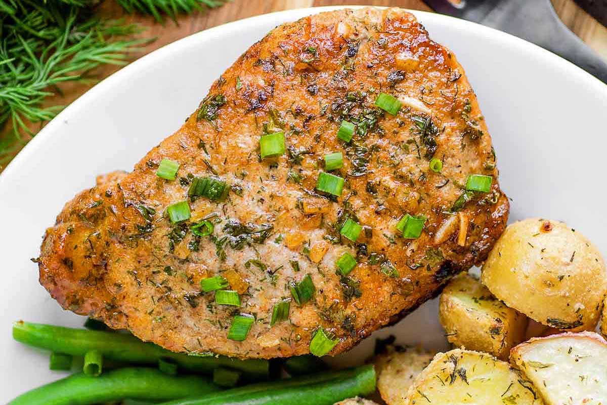 Grilled pork chop recipes garnished with herbs, served with roasted potatoes and green beans on a white plate.