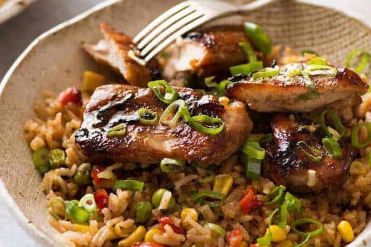 Plate of grilled chicken thighs on a bed of fried rice using frozen vegetables, garnished with chopped green onions, served with a fork.