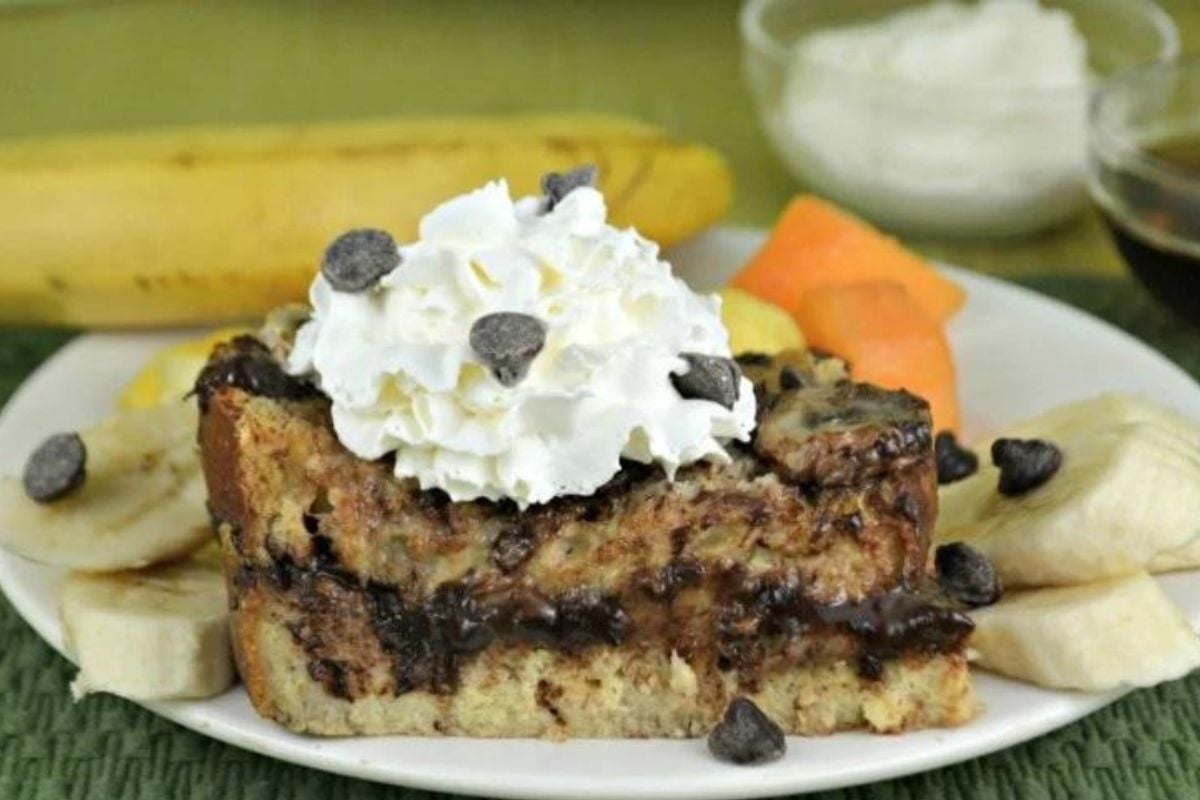 A slice of chocolate chip bread pudding topped with whipped cream and caramel, served with banana slices and a scoop of melon on the side.