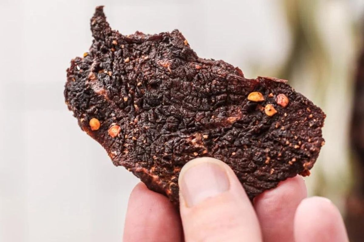 A close-up of a hand holding a piece of smoked beef jerky showing detailed texture and visible seasoning.