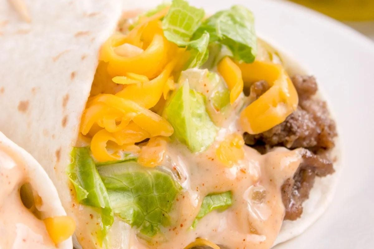 Soft taco with ground beef, shredded cheddar cheese, lettuce, and creamy sauce on a white plate—perfect for fans of classic ketchup recipes.