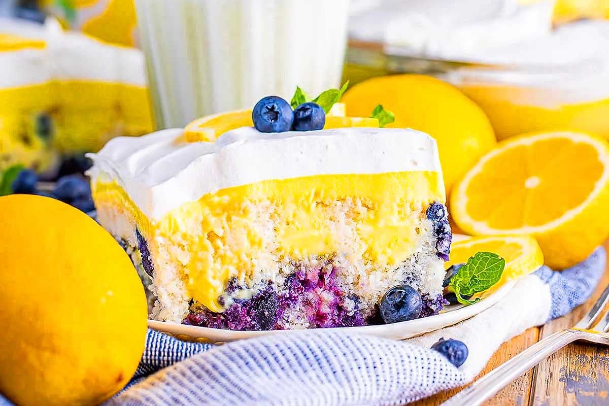 A slice of lemon blueberry cake made with pudding mix, garnished with fresh blueberries and mint on a plate with whole lemons in the background.