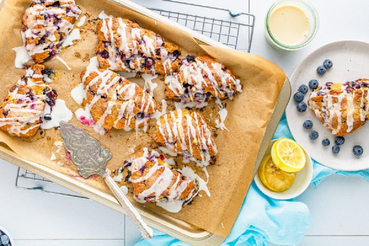 Freshly baked lemon blueberry scones with glaze drizzle on a baking tray, served with side lemons and blueberries from our collection of blueberry recipes.