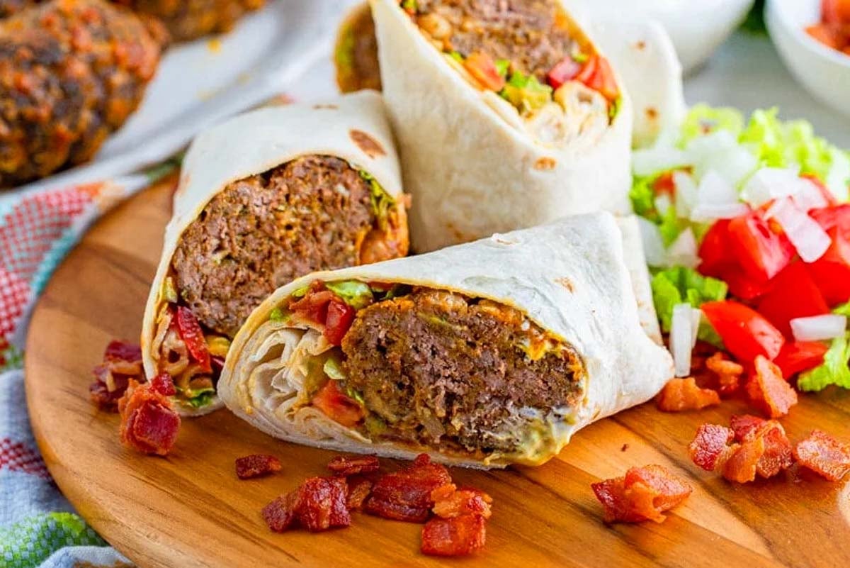 Two halves of a burrito-inspired meatloaf sandwich wrap with lettuce and tomatoes on a wooden board, garnished with diced tomatoes and bacon bits.