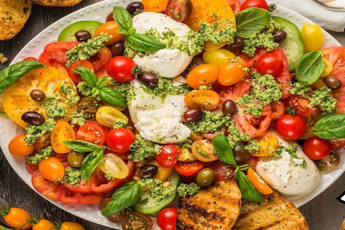 A plate of sliced tomatoes, burrata cheese, olives and a pesto dressing.