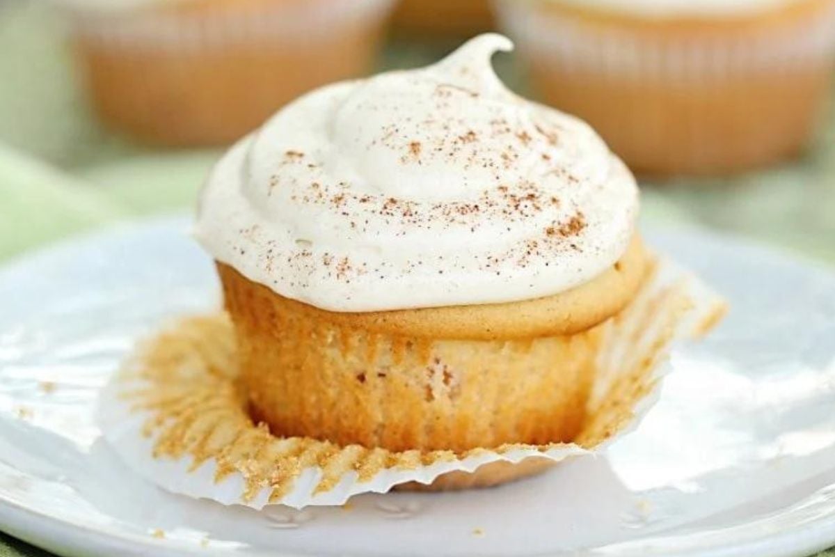 A vanilla cupcake with caramel frosting and a sprinkle of cinnamon on top, unwrapped and placed on a white plate.