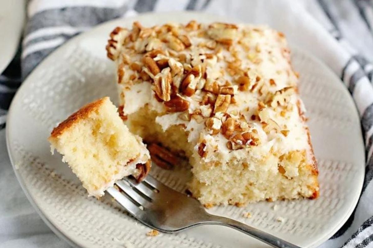 A piece of frosted caramel cake with chopped nuts on top, with one bite taken out, on a white plate with a fork.