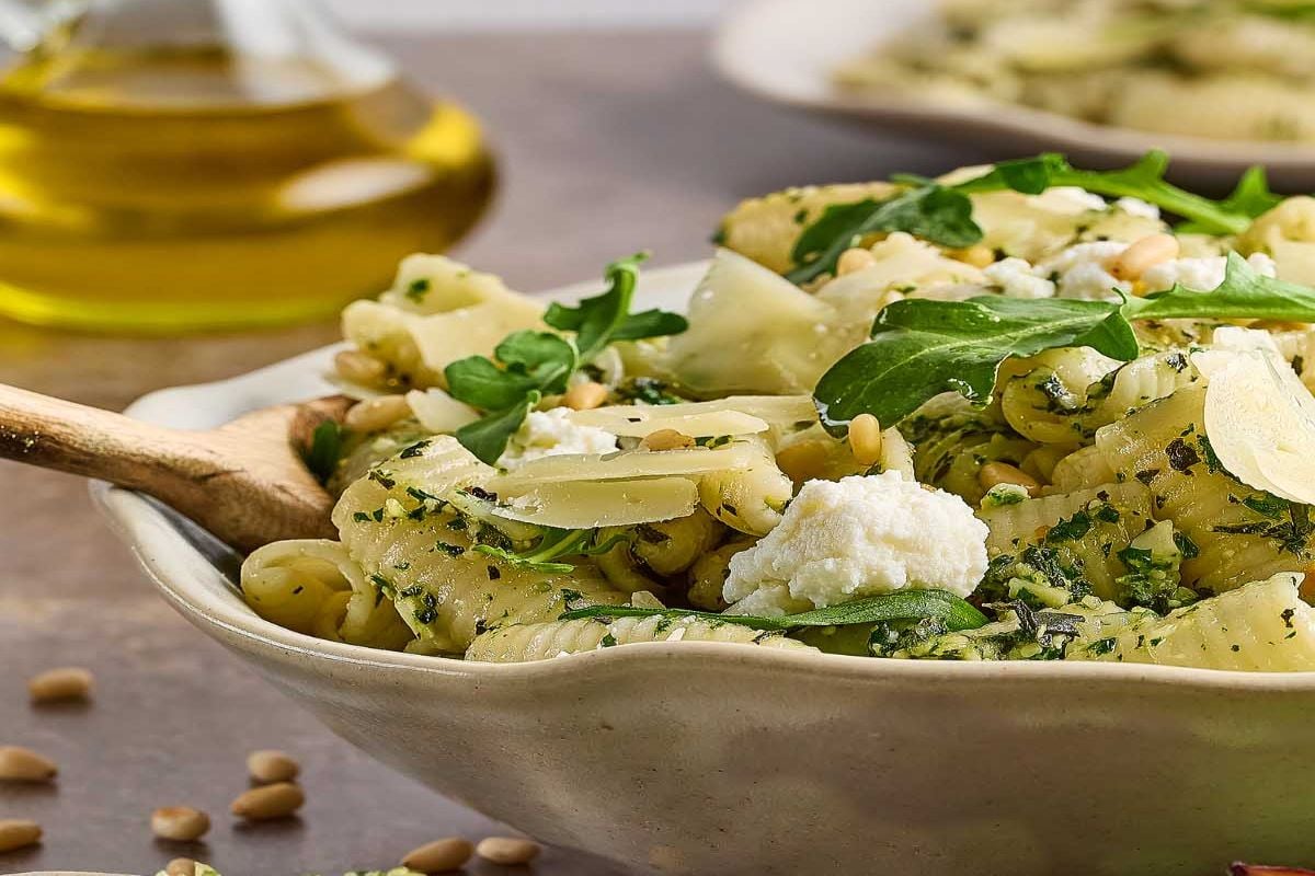 A bowl of pasta with pesto, ricotta, arugula, pine nuts, and parmesan—perfect for those seeking delicious pesto recipes. Olive oil sits in the background.