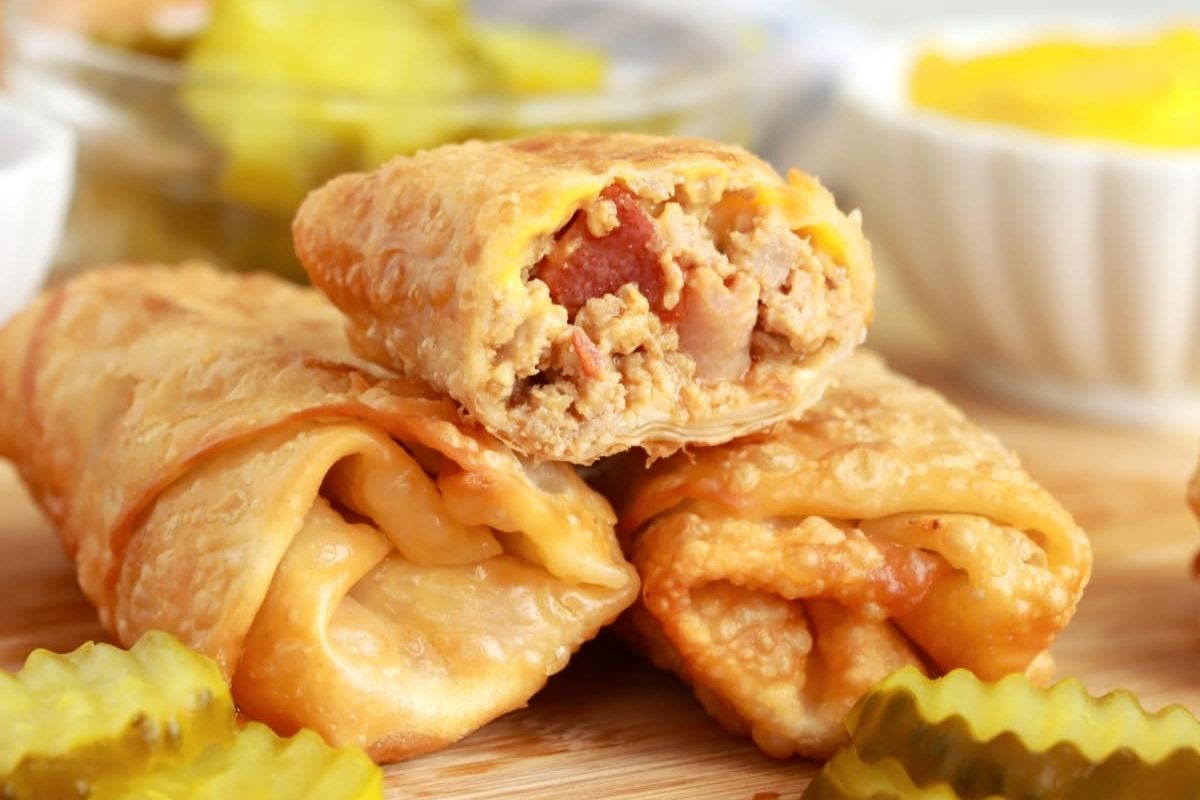 Close-up of crispy cheeseburger egg rolls on a wooden board, with pickle slices in the foreground—perfect for dipping and pairing with your favorite ketchup recipes.