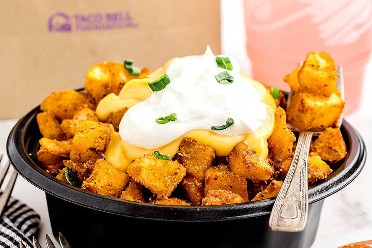 A bowl of seasoned potatoes topped with melted cheese and a dollop of sour cream, garnished with green onions, inspired by Taco Bell copycat recipes, on a table with a drink in the background