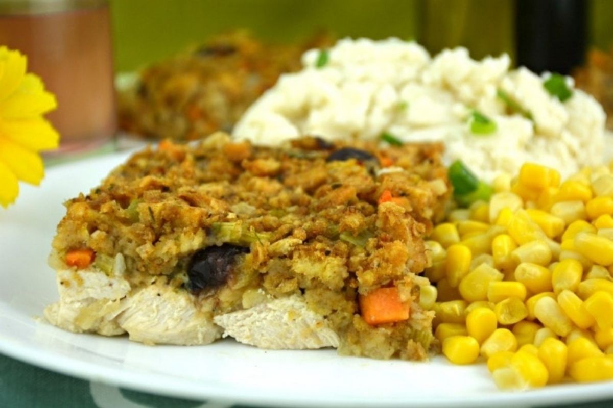 A plate of traditional food featuring a slice of chicken, stuffing, mashed potatoes with gravy, and corn.