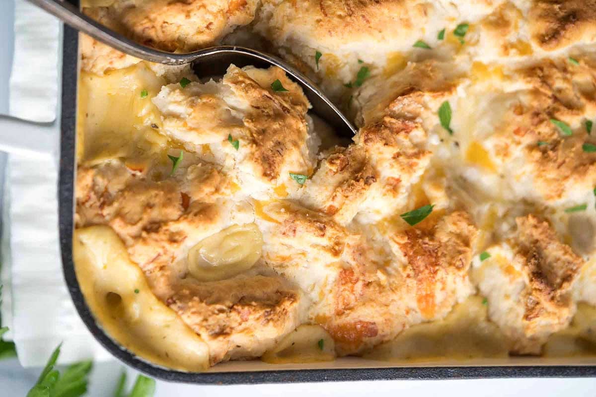 A spoon serving a portion of creamy chicken pot pie from a 9x13 baking dish with a flaky biscuit topping and visible chunks of chicken and vegetables.