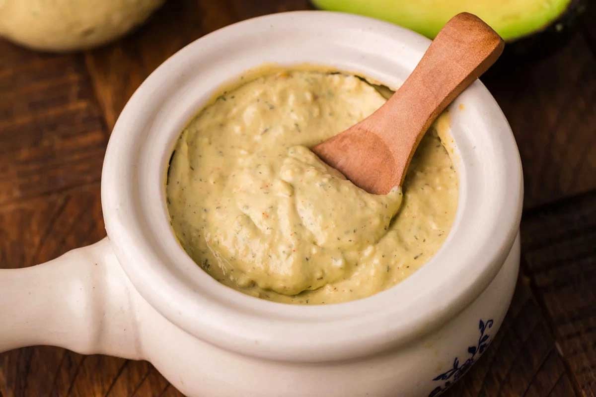 A bowl of creamy avocado dip, inspired by Taco Bell copycat recipes, with a wooden spoon, placed on a wooden table, with part of an avocado visible in the background.
