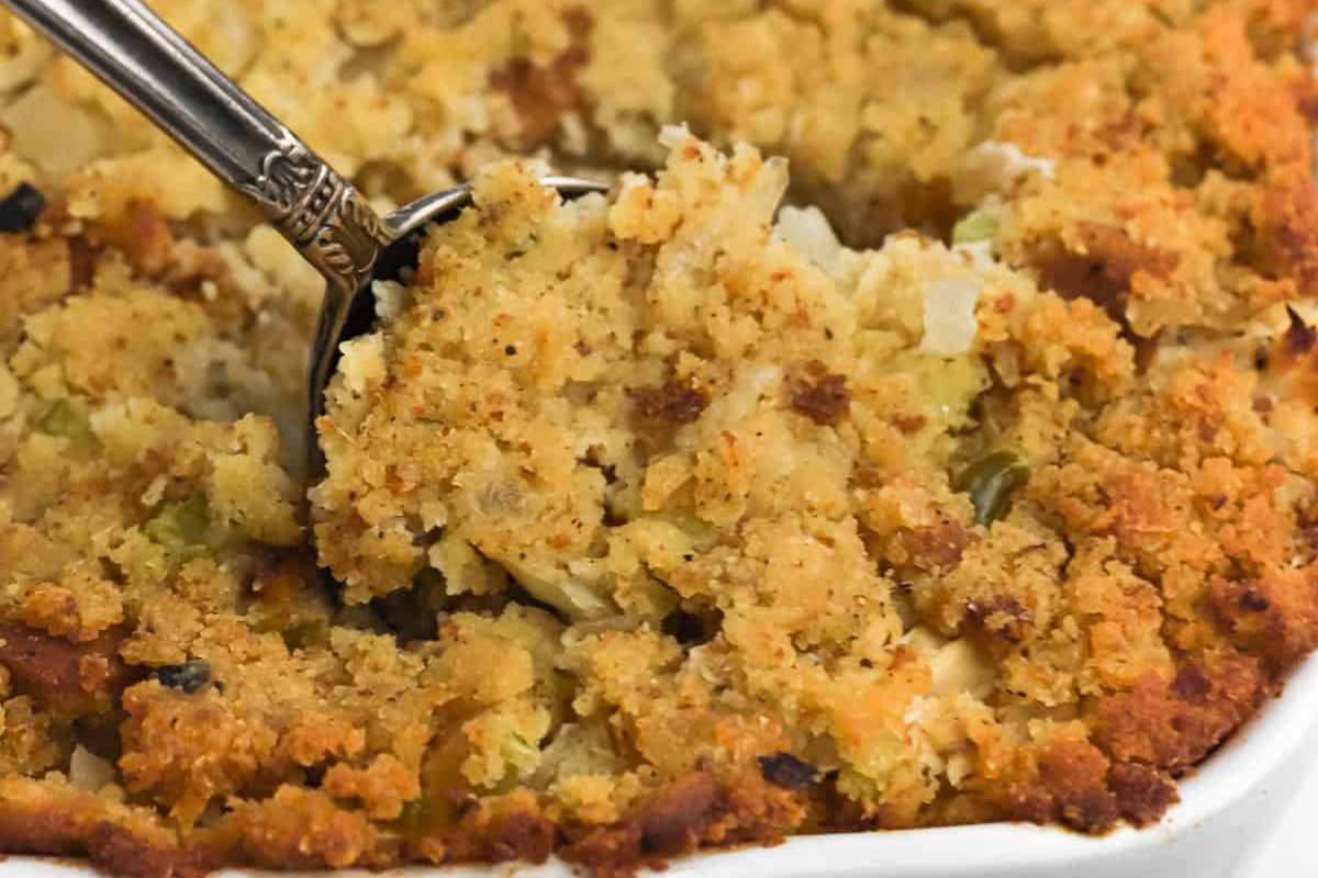 Close-up of a spoon scooping traditional bread stuffing with visible herbs and a can of cream soup, served in a white dish.