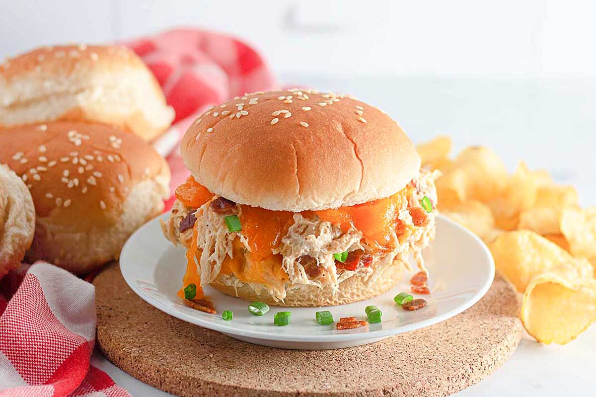 A slow cooker pulled pork sandwich on a sesame seed bun with side chips, garnished with green onions, on a white plate with a red napkin.