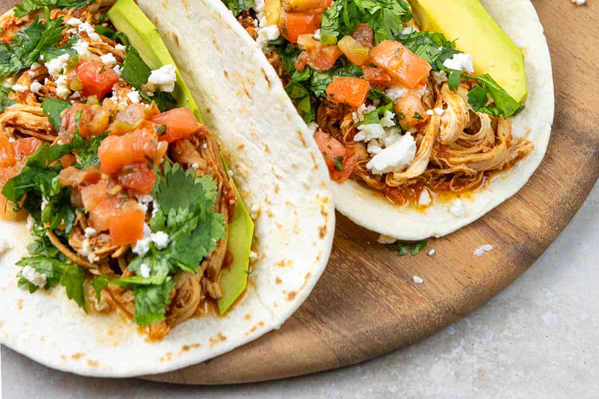 Two chicken tacos with avocado slices, fresh cilantro, and diced tomatoes prepared using slow cooker recipes and served on a wooden plate.