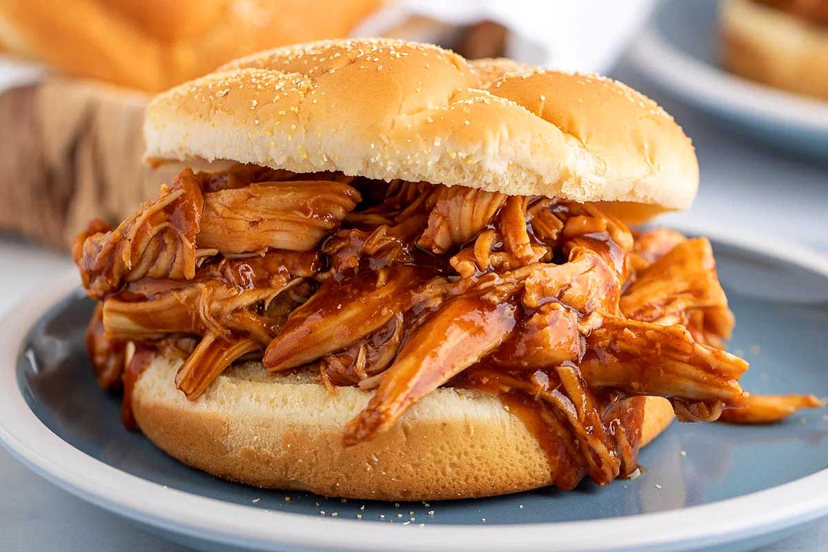 A slow cooker barbecued pulled chicken sandwich on a sesame bun, served on a white plate.