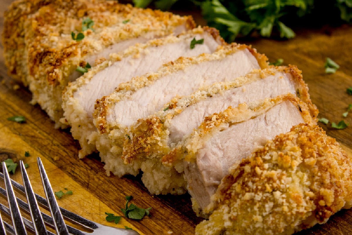 Sliced, breaded pork chop on a wooden cutting board, garnished with chopped parsley, with a fork alongside.
