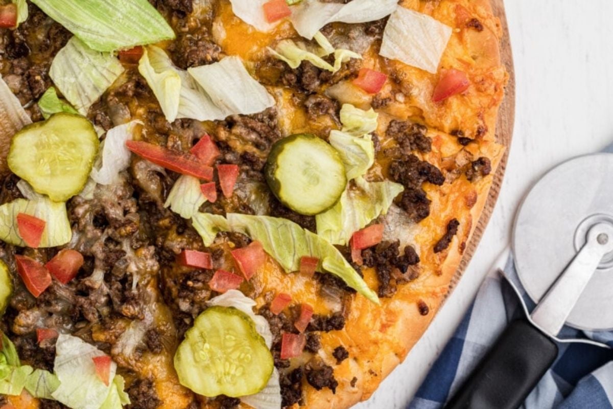 A Ground Beef Recipes-inspired pizza with ground beef, lettuce, tomatoes, onions, and pickles on a wooden pizza peel with a cutter.
