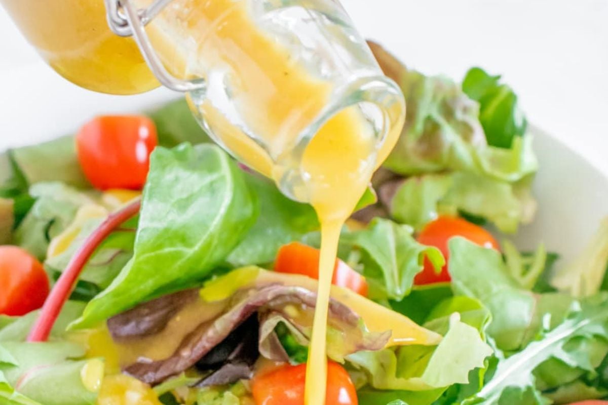 A bottle pours yellow salad dressing, inspired by Lemon Recipes, over a fresh salad with leafy greens and cherry tomatoes.