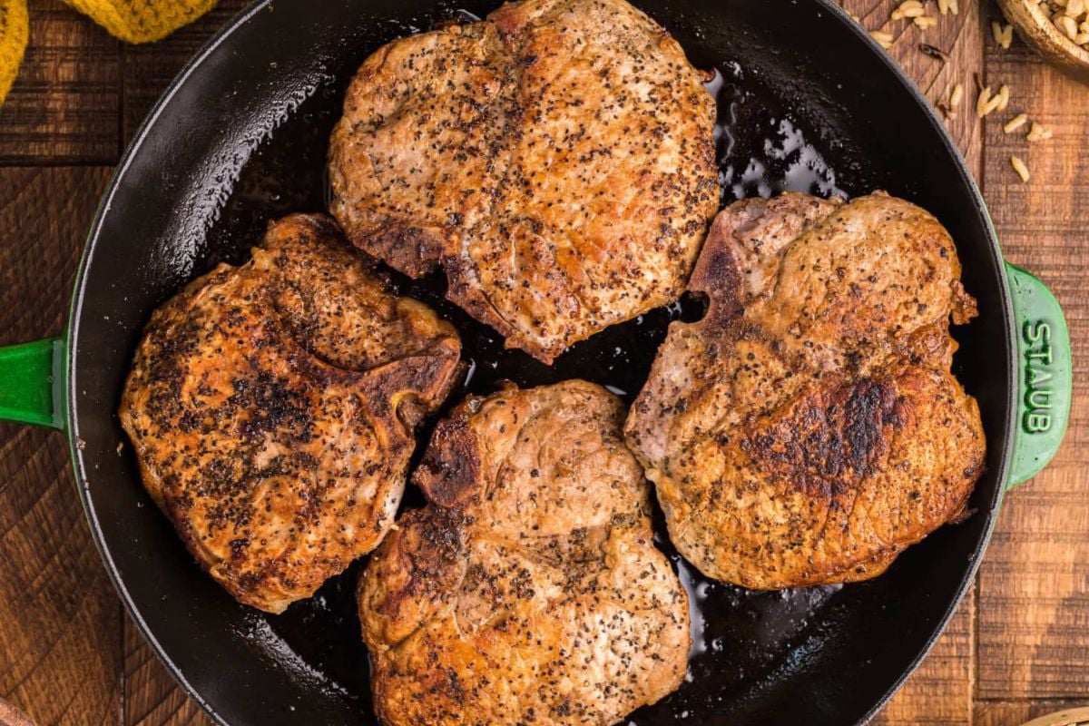 Four seasoned pork chop recipes in a cast iron skillet, viewed from above on a wooden table.