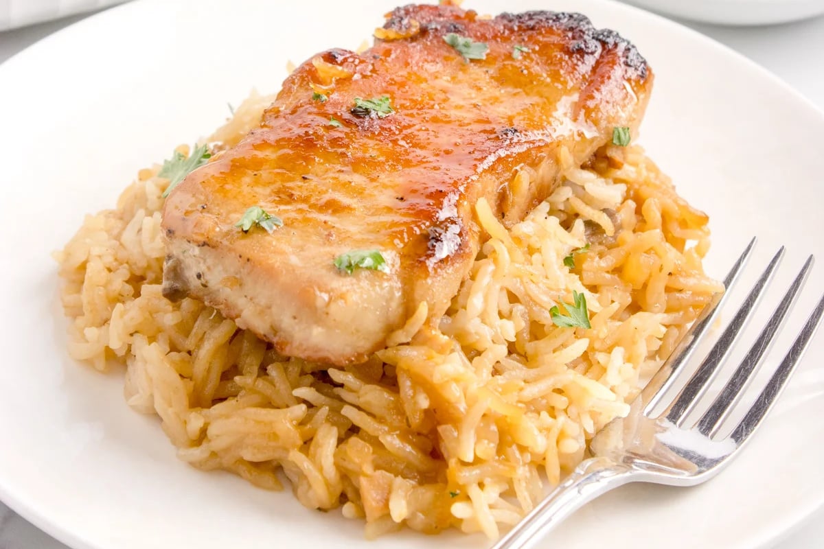 A glazed pork chop recipe served on a bed of seasoned rice garnished with herbs, on a white plate with a fork to the side.