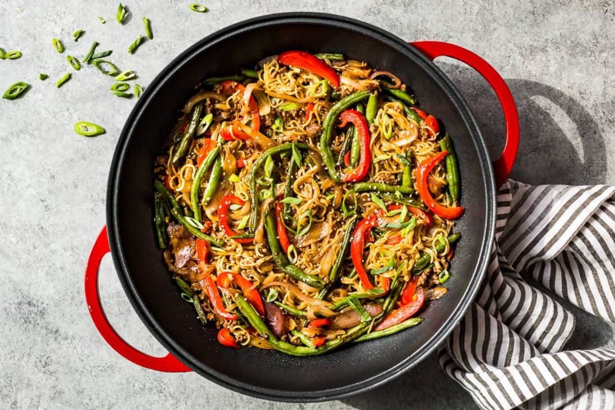 A black pan with stir-fried noodles, red peppers, green beans, and scallions on a gray surface, using frozen vegetables for added convenience.