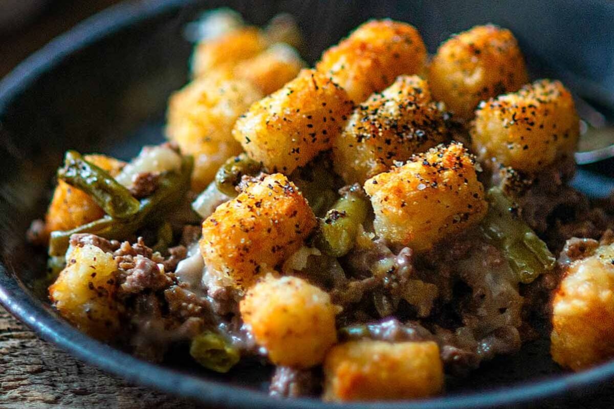A close-up of a dish containing seasoned tater tots, ground beef, a can of cream soup, and chopped green beans in a dark bowl.