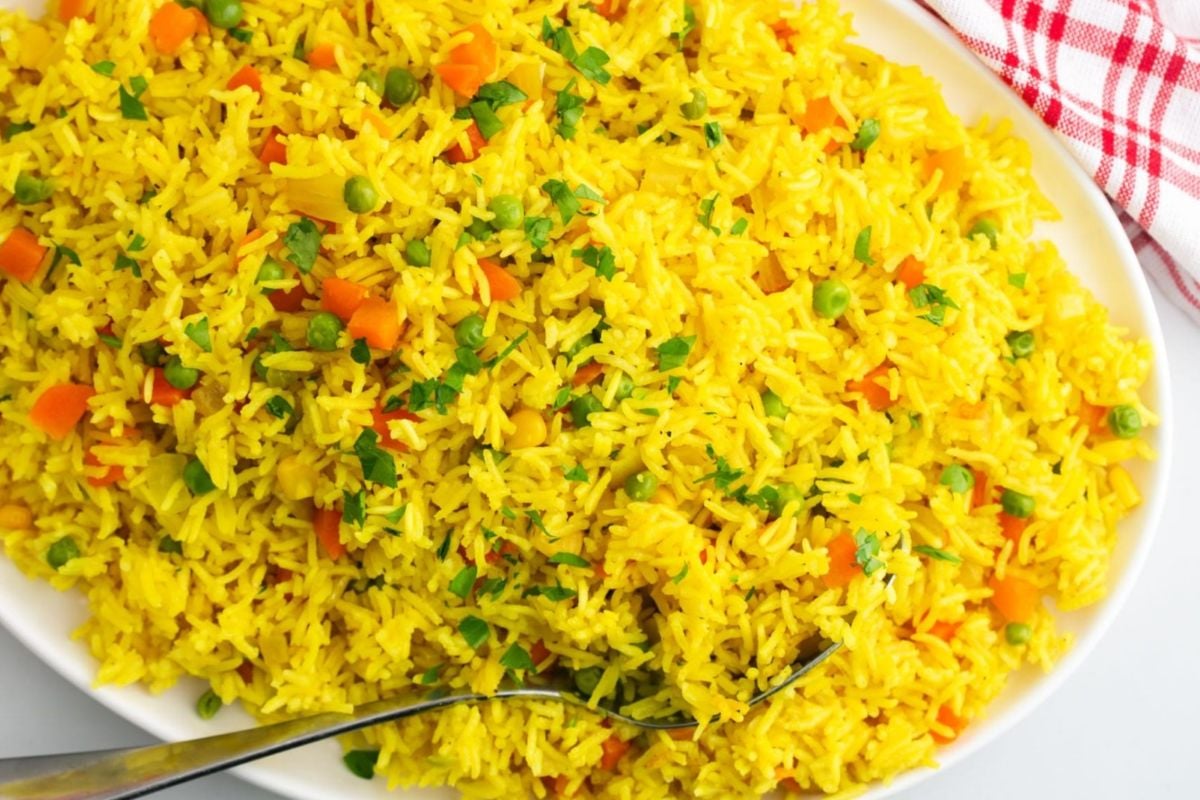 A large white plate filled with vibrant yellow vegetable fried rice made using frozen vegetables, garnished with chopped herbs, served with a fork on the side.