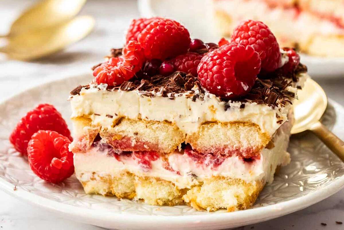 A slice of creamy raspberry tiramisu garnished with whole raspberries and chocolate shavings, served on an ornate white plate.