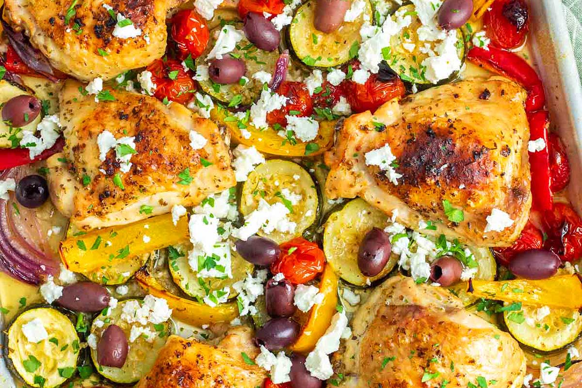Roasted chicken thighs surrounded by slices of zucchini, bell peppers, and tomatoes, garnished with feta cheese and olives in a sheet pan.