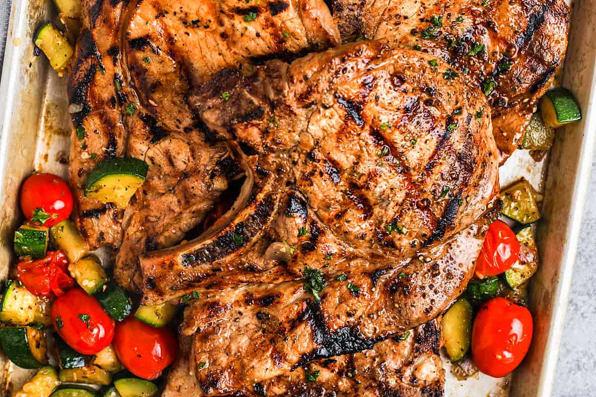 Grilled pork chop recipes garnished with herbs, surrounded by roasted tomatoes and zucchini in a cooking tray.